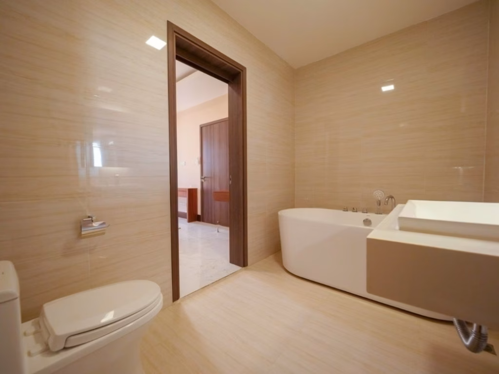 Modern bathroom with beige tiles, a white toilet, wall-mounted sink, and a freestanding bathtub in a stylish 2 bedroom condo. An open doorway leads to a hallway with brown doors and light-colored flooring.