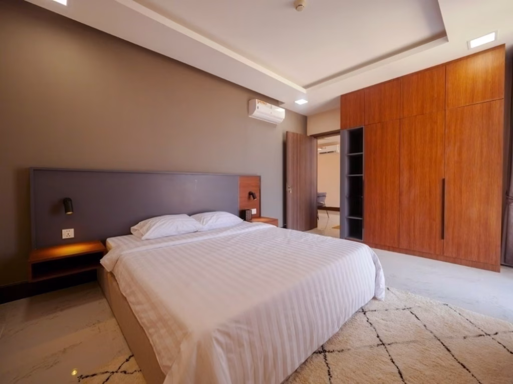 A modern 2 bedroom condo bedroom features a double bed with white striped bedding, wooden headboard, built-in nightstands, wooden wardrobe, beige walls, white tile floor, and a cream rug.