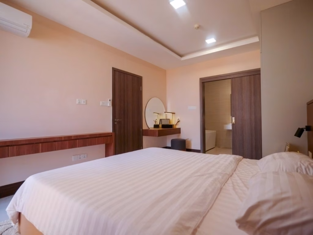 A modern 2 bedroom condo features a neatly made bed, a wooden desk, a round mirror with a stool, and an open door leading to a well-lit bathroom. The light peach walls and simple, elegant decor create a welcoming atmosphere.
