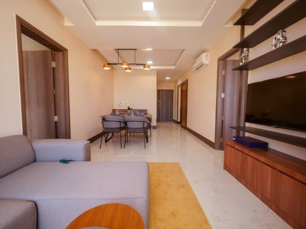 Modern living and dining area in a 2 bedroom condo with neutral decor, featuring a gray sofa, flat-screen TV on a wooden unit, dining table with chairs, ceiling lights, beige walls, and a tiled floor.