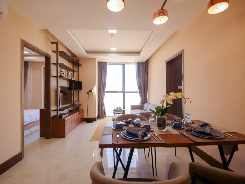 Modern open-plan living and dining area in this 2 bedroom condo, featuring a set dining table, cozy sofa, large window with curtains, TV unit, and shelves. Light walls and tiled floors create a bright, inviting space.