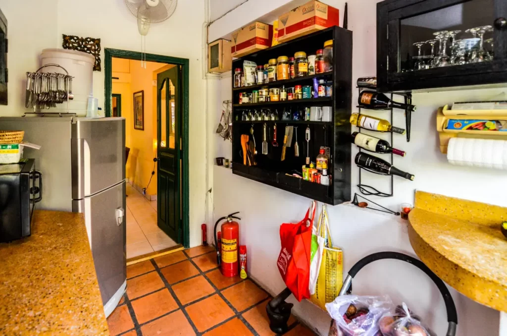 A cozy kitchen in Colonial Apartments Riverside with terracotta tile floors, open shelves holding spices, utensils, and wine bottles, a silver fridge, fire extinguisher, and a green door leading to another room.