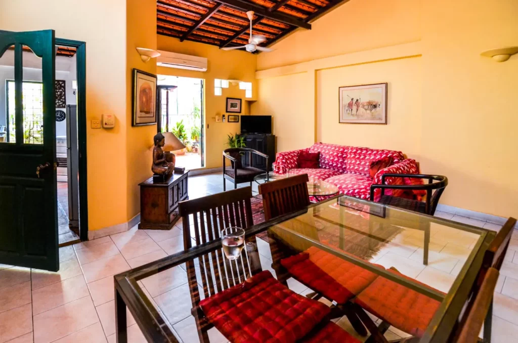 A bright living room in Colonial Apartments Riverside with yellow walls, a red patterned sofa, glass dining table with red-cushioned chairs, ceiling fan, wall paintings, and TV. Sunlight streams in through open doors and windows.