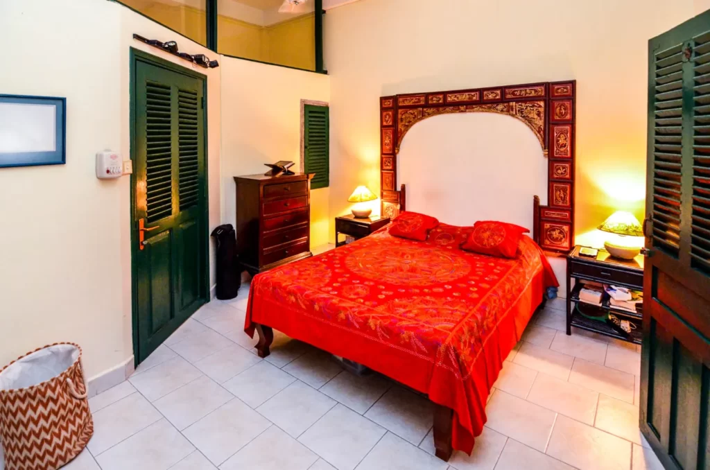 A cozy bedroom in colonial apartments riverside, featuring a double bed with a vibrant red and orange bedspread, bedside tables with lamps, a dark wooden dresser, wicker laundry basket, and green shuttered doors on a tiled floor.