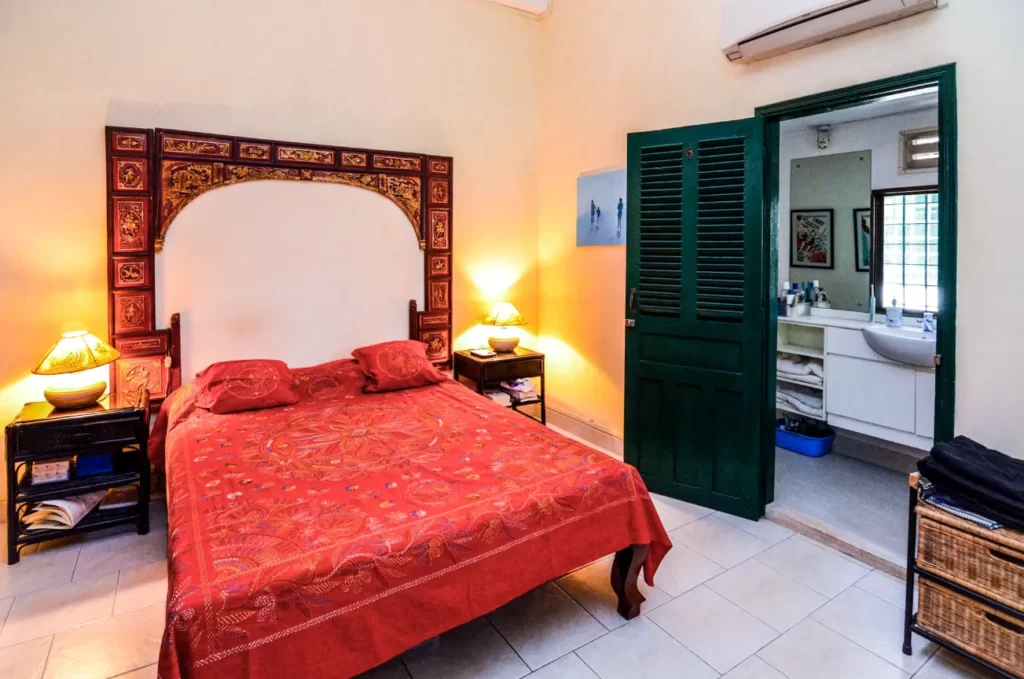 A cozy bedroom in Colonial Apartments Riverside features a red-patterned bedspread, two bedside tables with lamps, a decorative headboard, and an open green door leading to a well-lit bathroom with sink and mirror.