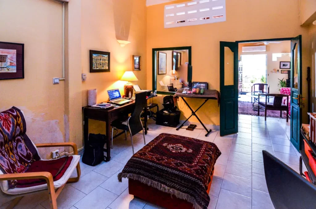 A cozy home office in Colonial Apartments Riverside features a desk, laptop, lamp, chair, patterned ottoman, and wall art. Green double doors open to another sunlit room. Warm yellow walls and tiled floors complete the inviting space.