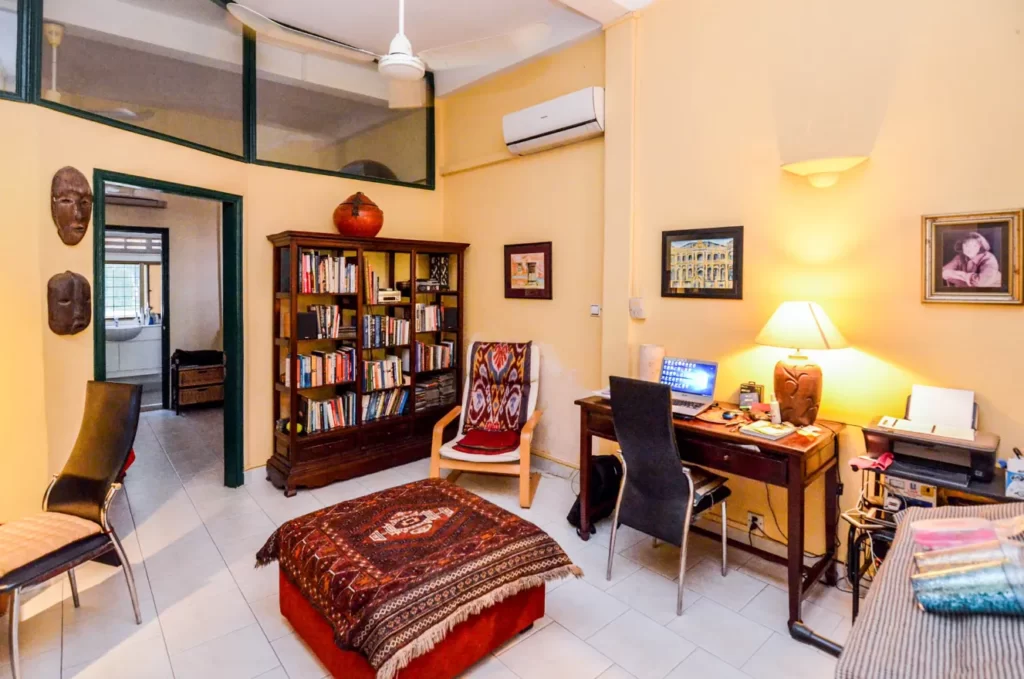 A cozy home office in Colonial Apartments Riverside features yellow walls, a wooden bookshelf filled with books, a desk with a laptop and lamp, a cushioned chair, two black chairs, a patterned ottoman, and stylish wall art.