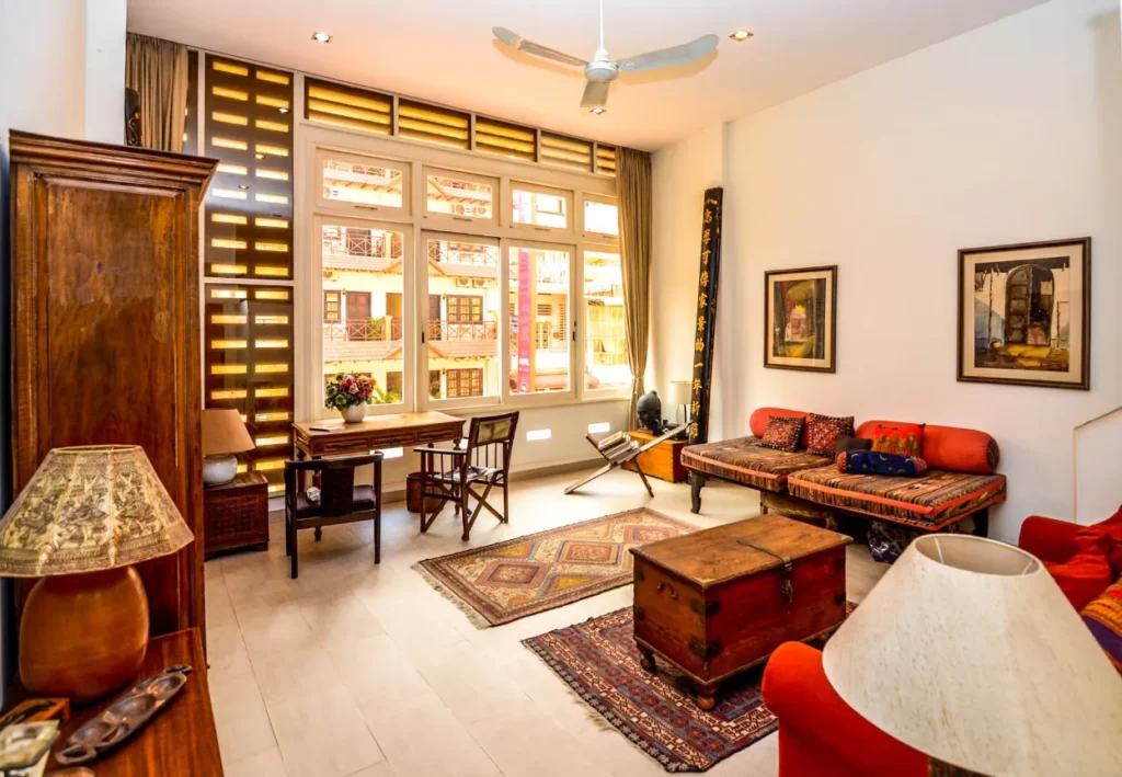 Bright living room with large windows at Colonial Apartments Riverside, wooden furniture, patterned rugs, a ceiling fan, vintage decor, and cozy seating with colorful cushions. Sunlight streams in, creating a warm and inviting atmosphere.