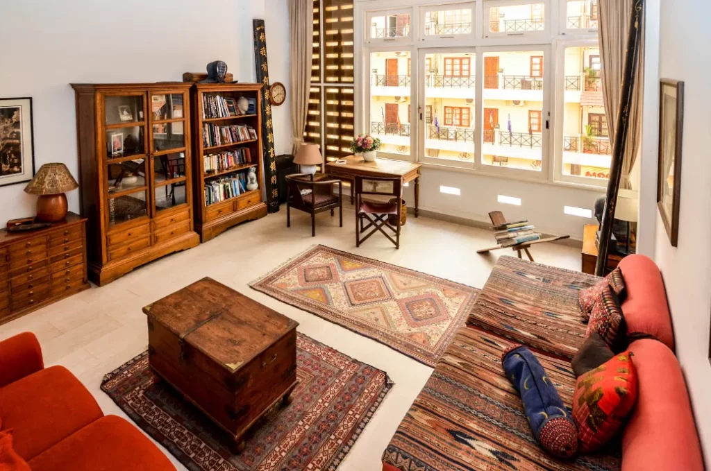 A cozy, sunlit living room in colonial apartments with a red sofa, patterned rugs, wooden furniture, bookshelves, and a writing desk by large windows. Colorful pillows brighten the space overlooking riverside balconies of an adjacent building.