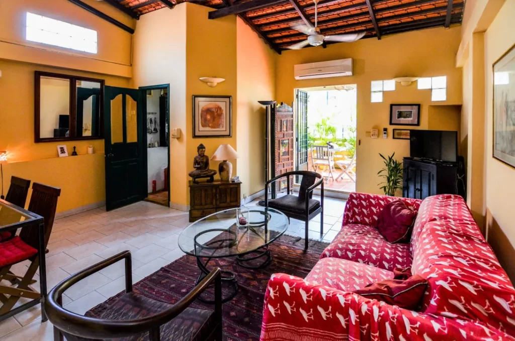 A cozy living room in Colonial Apartments Riverside features yellow walls, a red patterned sofa, glass coffee table, chairs, TV, and art decor. Sunlight streams in from open doors to a patio with outdoor seating and lush plants.
