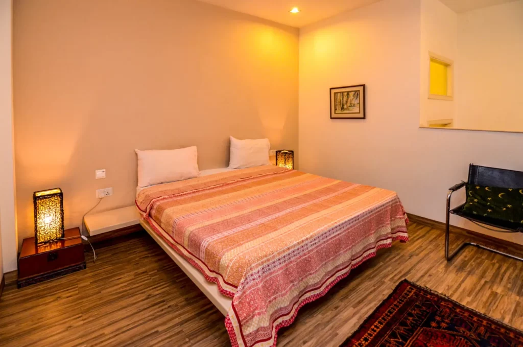 A cozy bedroom at Colonial Apartments Riverside features a double bed with a striped bedspread, two pillows, decorative lamps on either side, a wall painting, black chair, wooden flooring, and a red rug.
