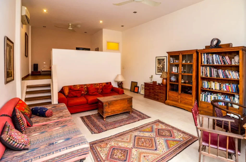A cozy living room at Colonial Apartments Riverside features colorful rugs, a red sofa, patterned bench, wooden coffee table, bookshelf filled with books, and framed art on white walls. Stairs and a partial wall separate the upper area.