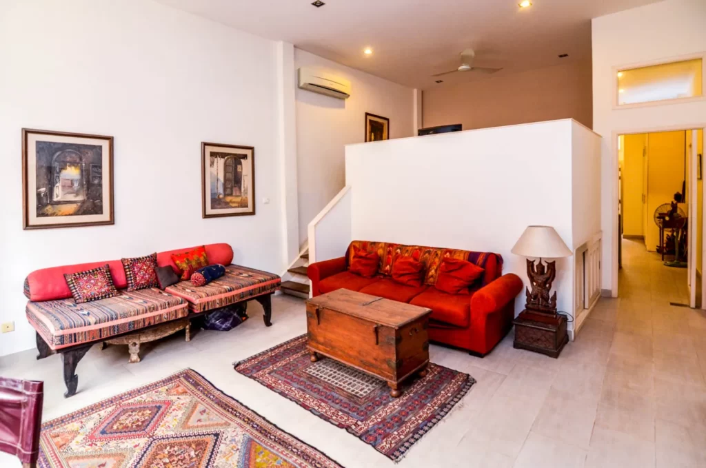 A cozy living room at Colonial Apartments Riverside features white walls, colorful patterned rugs, a red sofa, a low wooden coffee table, a patterned daybed, framed art, and a floor lamp in a modern, open-layout space with warm lighting.