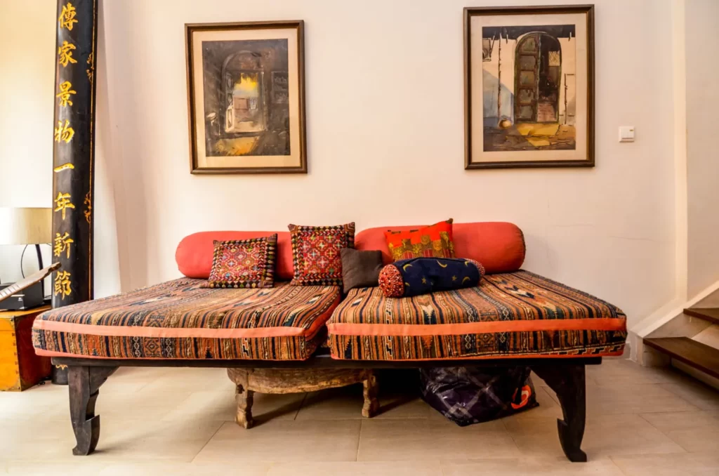 A cozy seating area in colonial apartments riverside features two patterned cushions, colorful pillows, and a low wooden bench beneath framed paintings on a white wall. A stairway is visible on the right.