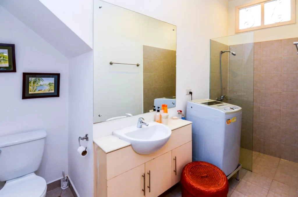 A bathroom in Colonial Apartments Riverside features a toilet, sink vanity, mirror, washing machine, and glass-enclosed shower. Framed pictures adorn the wall, with a red round stool placed next to the washing machine for convenience.