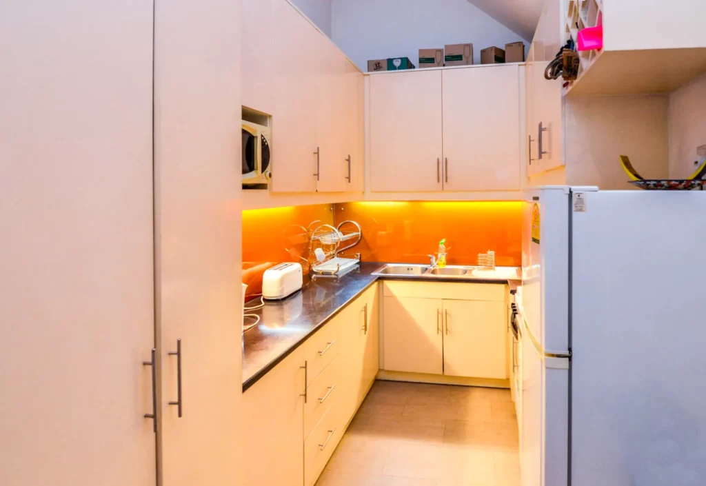 Modern kitchen at Colonial Apartments Riverside with cream cabinets, stainless steel countertops, orange under-cabinet lighting, toaster, dish rack, microwave, and white refrigerator. Storage boxes are neatly placed above the cabinets.
