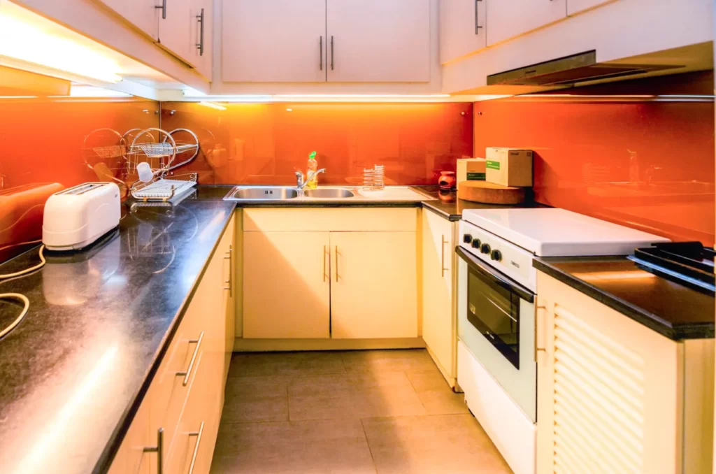 A modern kitchen at Colonial Apartments Riverside features white cabinets, black countertops, an orange backsplash, a white oven, toaster, dish rack, and a sink with a faucet under bright lighting.