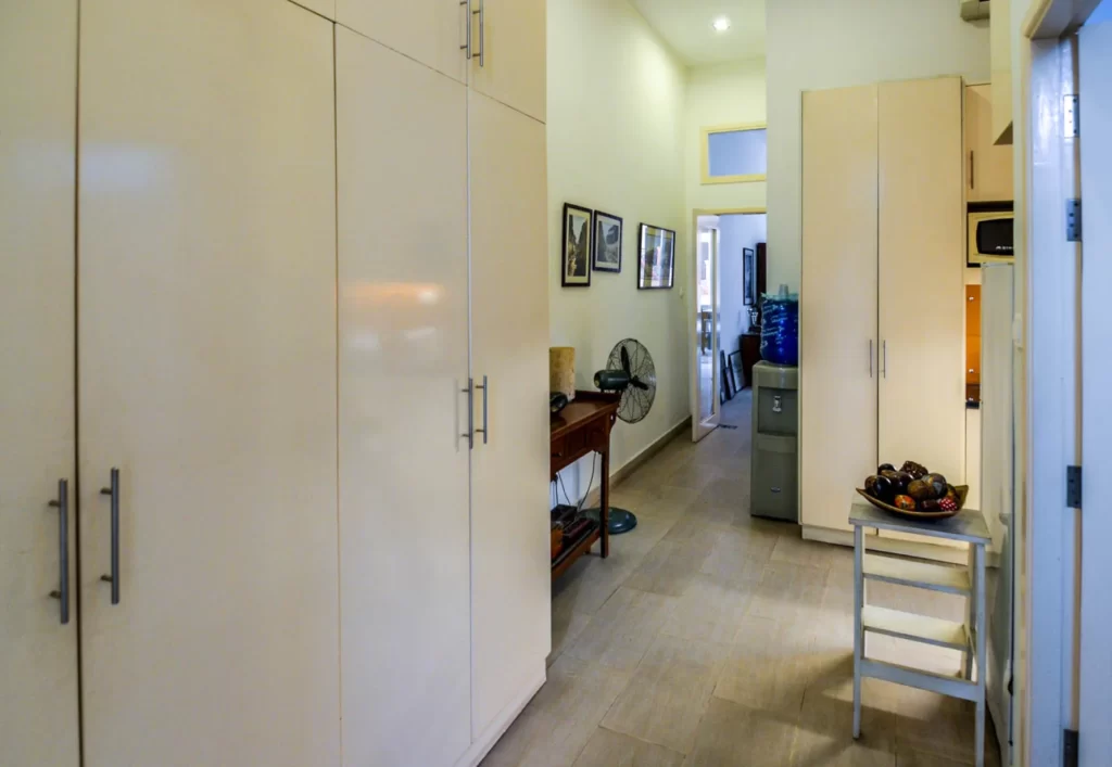 A hallway in Colonial Apartments Riverside features beige cabinets on both sides, a small table with a fruit bowl and stool, framed pictures on the wall, a fan, and a water dispenser in the background.