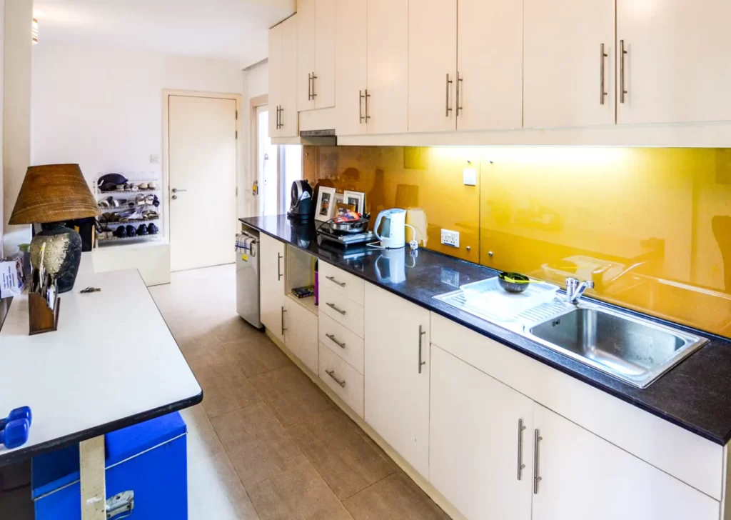 A modern kitchen at Colonial Apartments Riverside features white cabinets, black countertops, a stainless steel sink, yellow backsplash, and appliances like a kettle and toaster. A table with a lamp sits left, with shelves for shoes near the door.