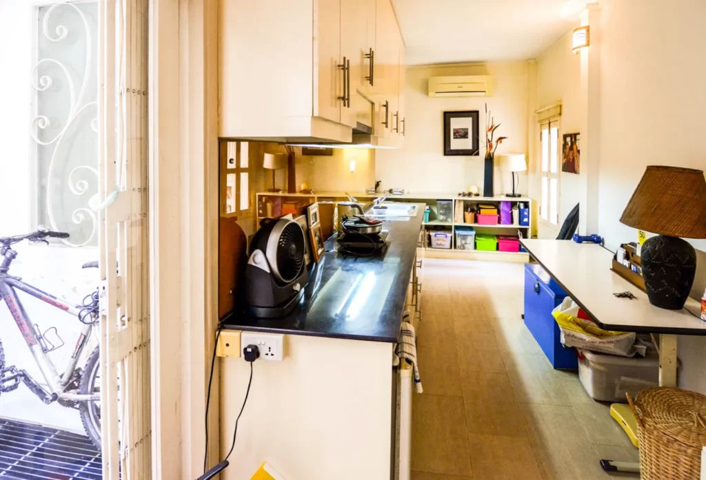 A bright, narrow kitchen at Colonial Apartments Riverside features white cabinets, dark countertops, and modern appliances. A bicycle by the door and colorful storage bins line the far wall beneath a window and air conditioner.