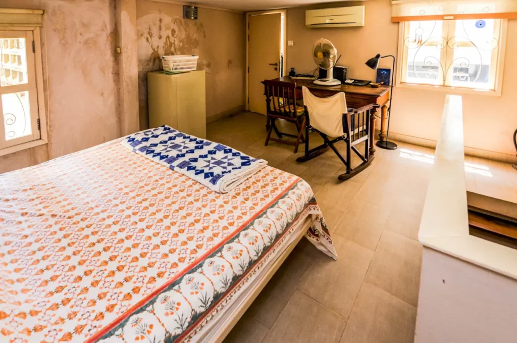 A cozy bedroom at Colonial Apartments Riverside features a double bed with a patterned orange and white bedspread, a blue and white blanket, a wooden desk and chair, a fan, computer, and large windows letting in natural light.