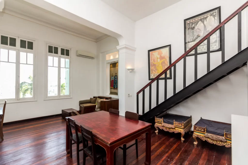 A bright living and dining room with white walls, dark wood floors, a wooden dining table with chairs, two traditional couches, framed art, and a black metal staircase with ornate seats beneath it.