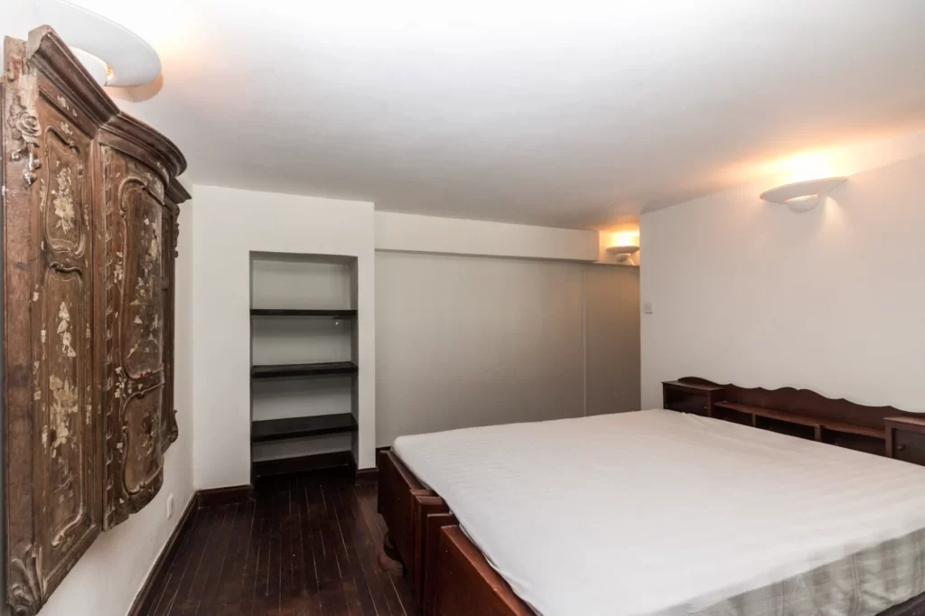 Minimalist bedroom with a large bed, dark wooden floor, ornate wooden wardrobe, built-in shelving, and white walls softly lit by wall-mounted lights.