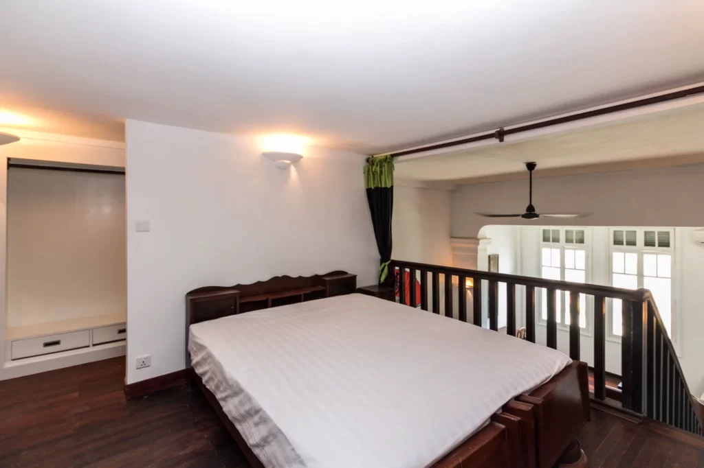 A loft bedroom with a large bed, dark wood flooring, white walls, built-in wardrobe, and a railing overlooking a lower level with tall windows and a ceiling fan.