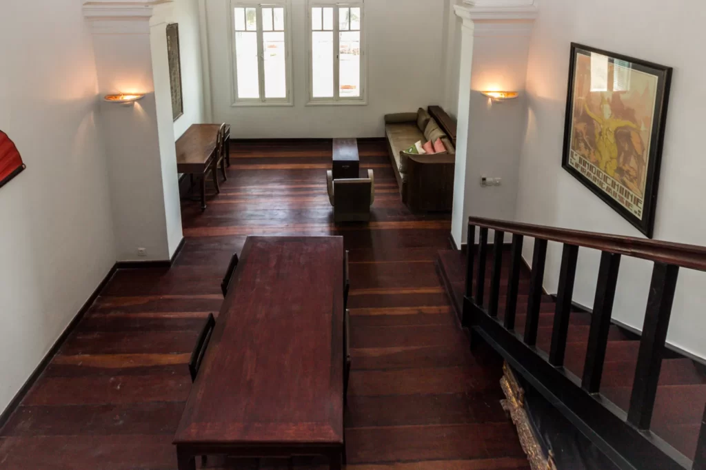 View from above of a spacious room with dark wooden floors, a dining table, a desk by the windows, a couch, framed art on white walls, and a staircase with black railings. Light streams in from the windows.