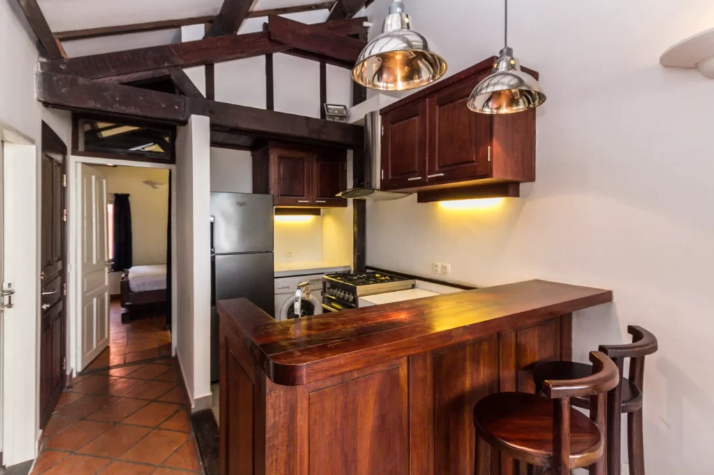 A cozy kitchen with dark wooden cabinets and a matching breakfast bar, two barstools, stainless steel appliances, pendant lights, and exposed wooden beams. A doorway leads to a bedroom in the background.