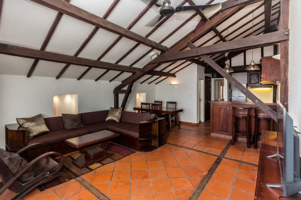A cozy living and dining area with exposed wooden beams, terracotta tile flooring, a brown sectional sofa, wooden dining table, and an open kitchen with dark wood cabinets and bar seating under a sloped ceiling.