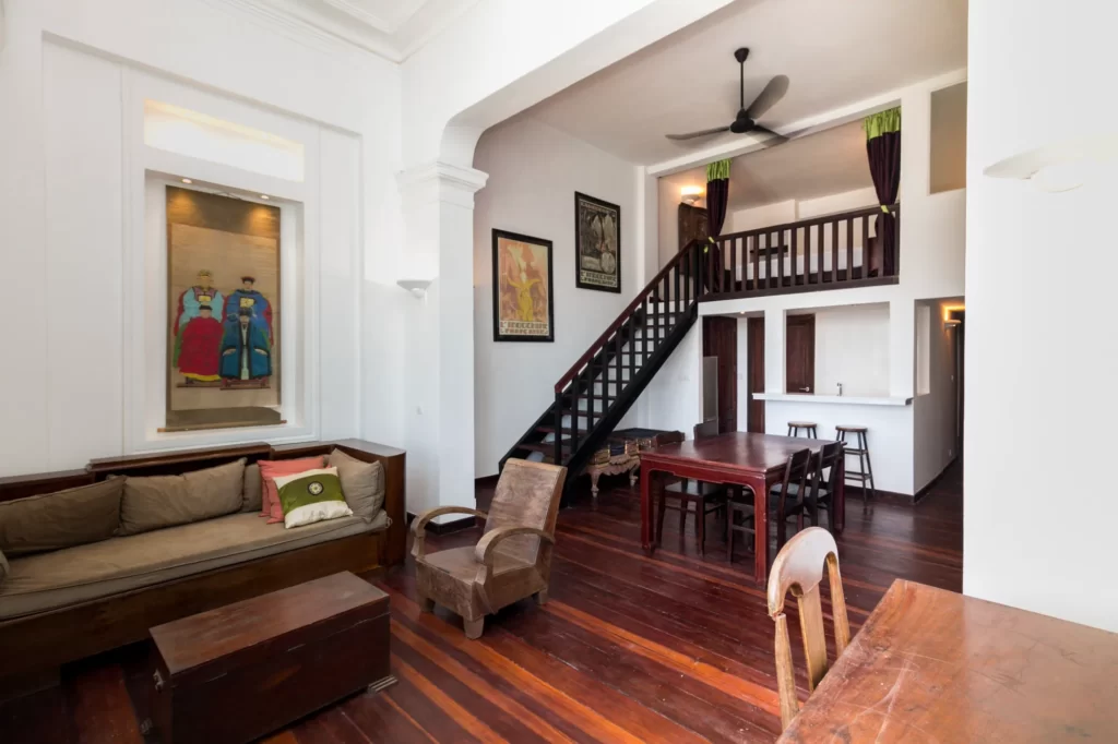 A spacious living area with wooden floors, a sofa, armchairs, dining table, art on the walls, and a loft with a staircase. The room features high ceilings, white walls, and a ceiling fan.