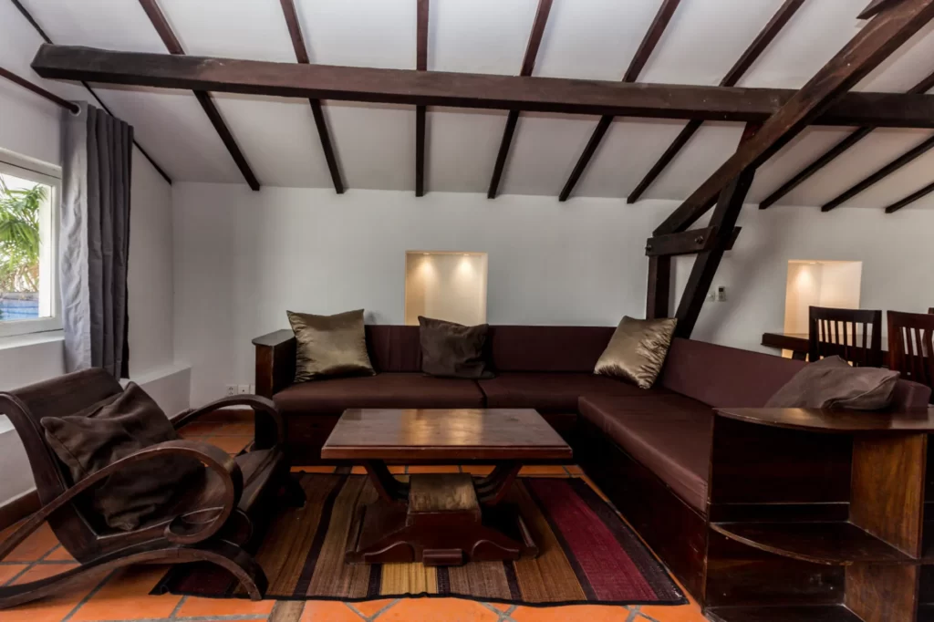 A cozy living room with dark wooden furniture, including a corner sofa, coffee table, and chair, set on a patterned rug. Slanted ceiling with exposed beams and light streaming in from a window on the left.