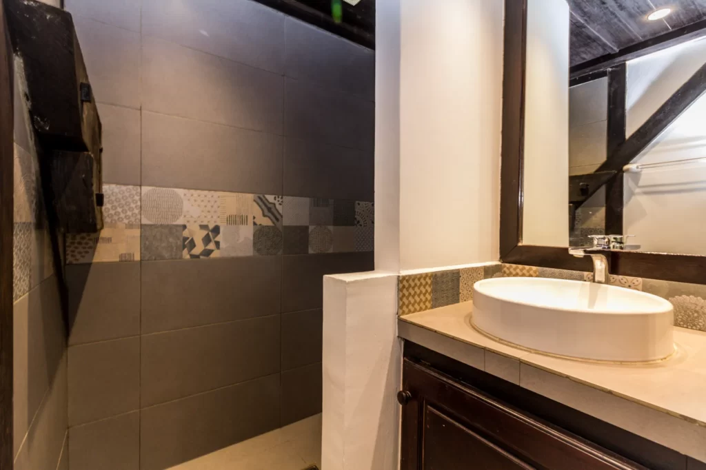 Modern bathroom with dark tiled walls, a patterned accent strip, a white vessel sink on a countertop, a large mirror, and a partially open walk-in shower.