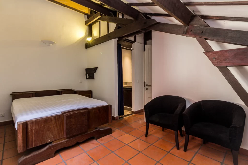 A cozy bedroom with a wooden bed, two black armchairs, terracotta tile flooring, white walls, exposed dark wooden beams, and an open doorway leading to another room.