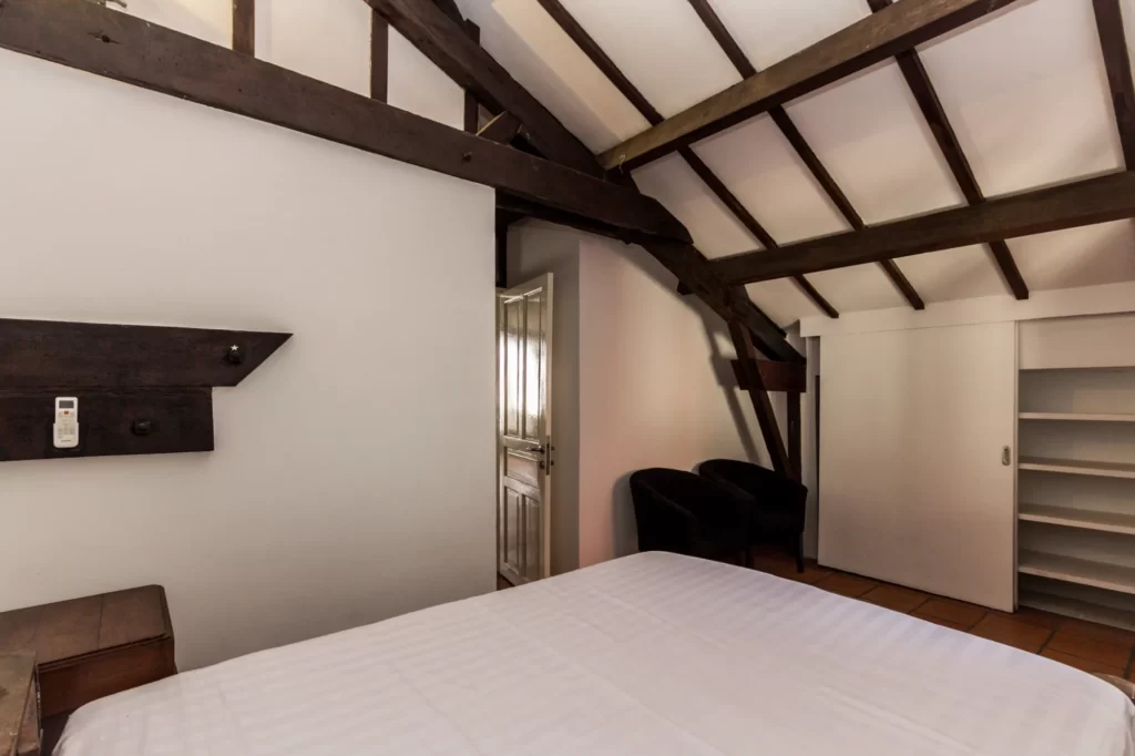 Attic bedroom with exposed dark wooden beams, a white bed, two black chairs, a shelf unit with sliding doors, and a partially open door letting in daylight.