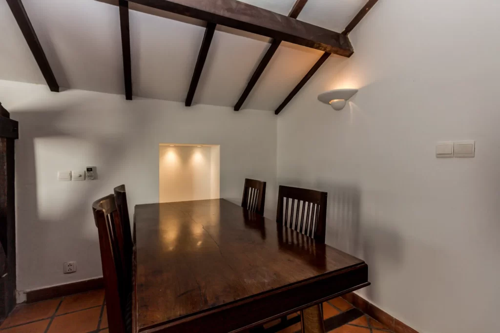 A wooden dining table with four chairs sits beneath a slanted ceiling with exposed wooden beams; a white wall and two recessed lights are in the background.
