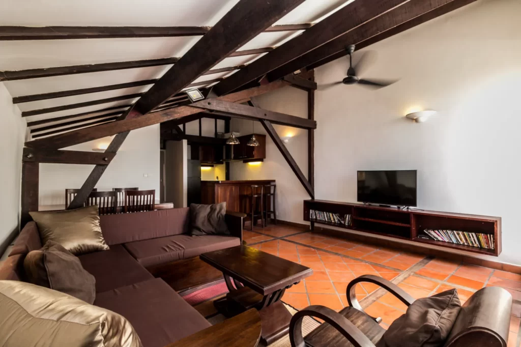 A cozy living room with wooden beams, brown sofas, a TV on a low stand, bookshelves, and a ceiling fan. The space has terracotta tile flooring and an open kitchen with bar stools in the background.
