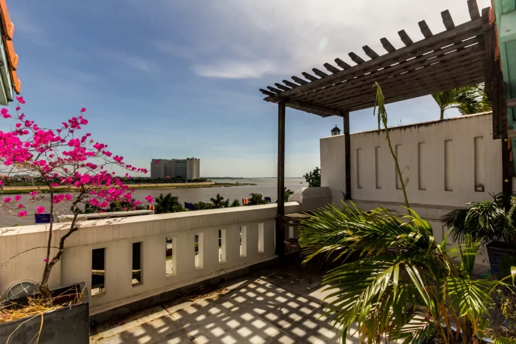 A rooftop terrace with a wooden pergola overlooks a river, city buildings, and a clear blue sky. Pink bougainvillea and green palm plants add vibrant color to the sunlit scene.
