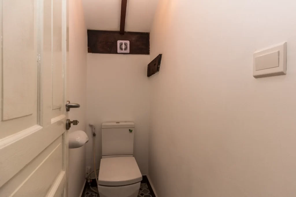 A small, narrow bathroom with white walls, a closed white door, a toilet, and a wooden beam on the ceiling. There is a light switch on the wall and an exposed patch on the right wall.