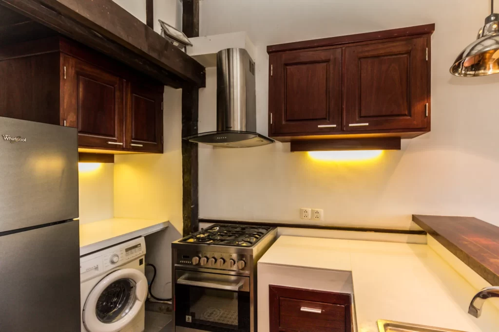 Modern kitchen with dark wood cabinets, a stainless steel gas stove, a range hood, under-cabinet lighting, a refrigerator, and a washing machine next to the counter on a white and wood-accented background.