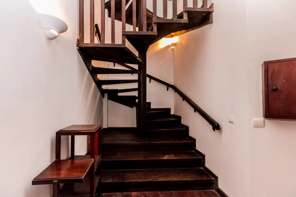 Wooden staircase with dark brown steps and railing, turning at a right angle in a white-walled interior. A small wooden table with shelves is placed near the bottom of the stairs. Wall-mounted lights provide illumination.
