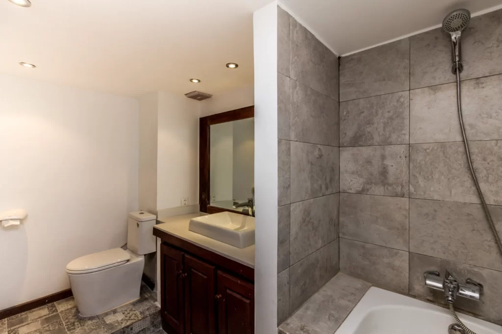 Modern bathroom with a tiled bathtub and shower on the right, a wooden vanity with a sink and mirror in the center, and a toilet on the left, all under recessed ceiling lights.