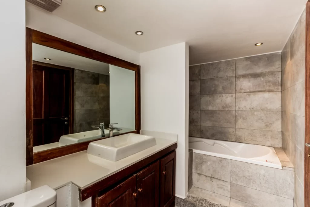 Modern bathroom with a large mirror above a double sink vanity, wooden cabinets below, and a bathtub with tiled walls in the background. The space features recessed lighting and neutral tones.