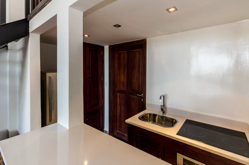 A compact modern kitchen with dark wood cabinets, a stainless steel sink, an electric cooktop, white countertops, and a closed wooden door. Recessed ceiling lights brighten the space.
