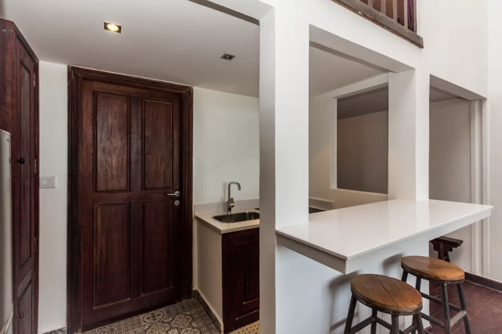 A small kitchen with dark wooden cabinets and door, a single sink, patterned floor tiles, and a white countertop bar with two wooden stools. The area has white walls and open shelving above the bar.
