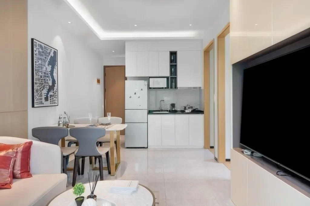 Modern, compact studio apartment for comfortable living, featuring a small kitchen, dining table and chairs, a sofa with red pillows, wall art, and a large flat-screen TV. The bright space boasts light-colored decor and contemporary furnishings.