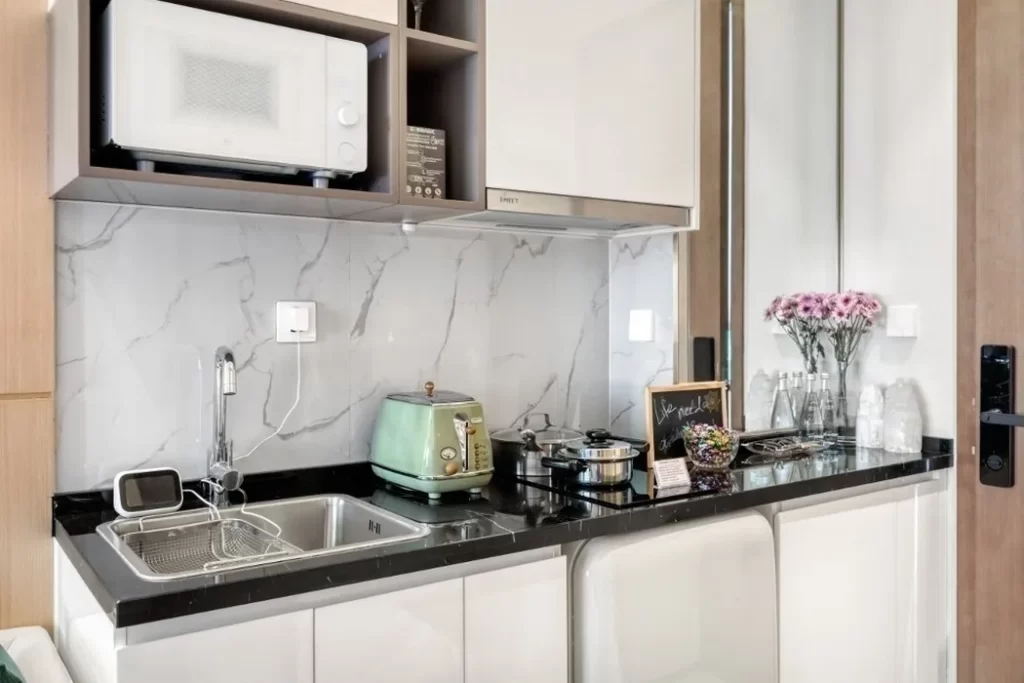 Modern kitchen in a studio apartment for rent, featuring a marble backsplash, microwave, green toaster, sink, stove, flower vase, glasses, and a small chalkboard on a tidy black countertop. The space is well-organized and inviting.