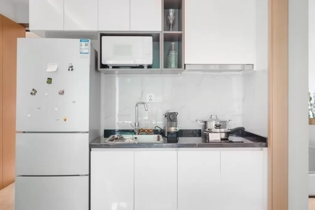 A modern, compact kitchen in a studio apartment for easy living, featuring white cabinets, a refrigerator with magnets, a microwave, sink, coffee maker, and pot on the stove set against a white marble backsplash.