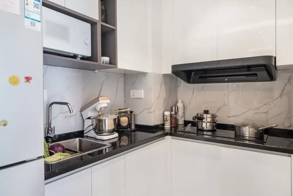 Modern kitchen in a studio apartment for convenient living, featuring white cabinets, a black countertop, and a marble-patterned backsplash. Appliances include a microwave, stand mixer, pots on the stove, and a magnet-covered refrigerator. Vegetables fill the sink.